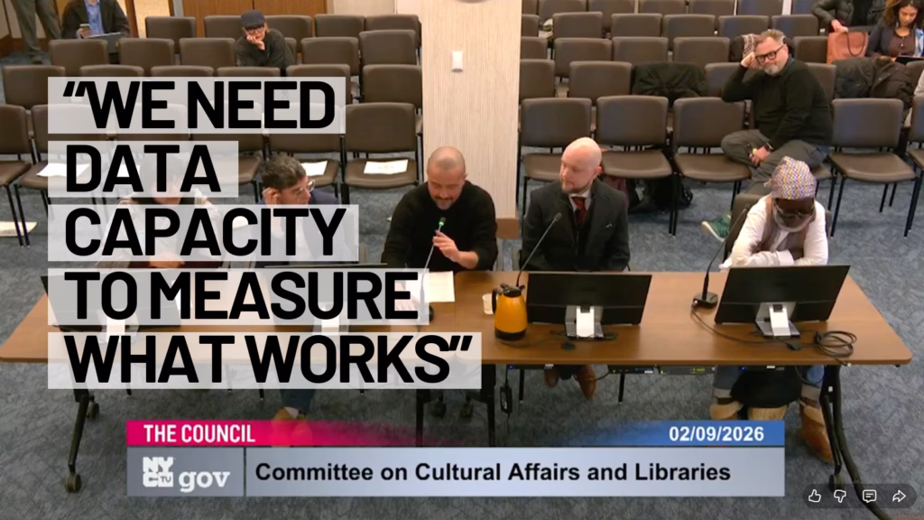 Wide shot of a New York City Council hearing room. Four people sit at a table with microphones and monitors; one person is speaking into a microphone. Rows of mostly empty chairs are behind them. Large overlaid text reads, ‘We need data capacity to measure what works.’ A banner at the bottom shows ‘The Council,’ ‘Committee on Cultural Affairs and Libraries,’ and the date 02/09/2026.